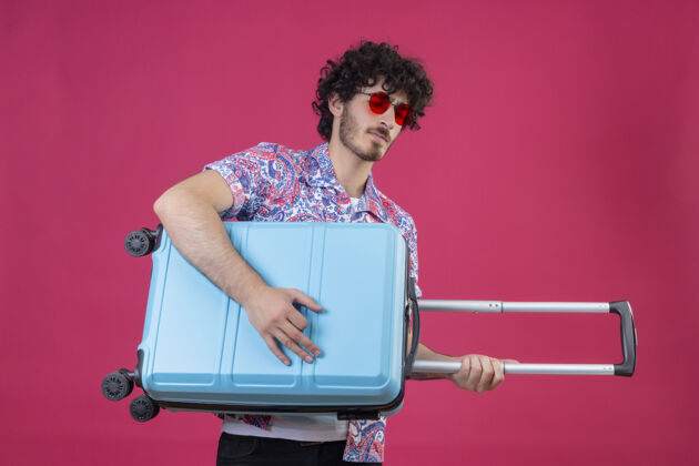 男人自信的年轻帅气卷发旅行者戴着太阳镜 用手提箱当吉他在孤立的粉色空间穿旅行者手提箱