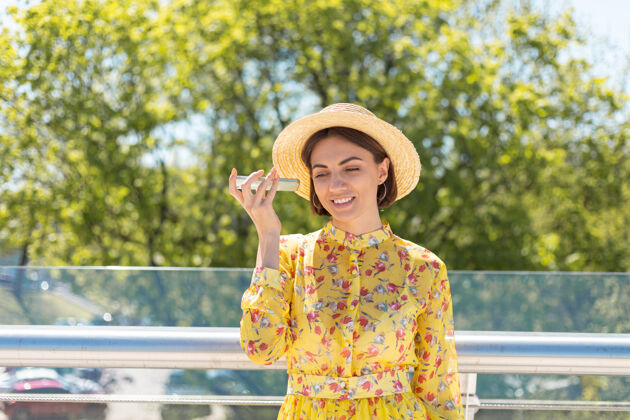 城市穿着黄色夏装戴着帽子的女人的户外肖像在电话里听语音留言玩应用休闲