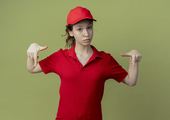 女孩令人印象深刻的年轻漂亮的送货女孩在红色制服和帽子指向下孤立的橄榄绿背景与复制空间背景指向绿色