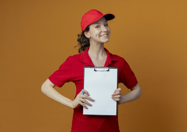 显示微笑着的年轻漂亮的送货女孩 穿着红色制服 戴着帽子 手里拿着钢笔 对着相机展示剪贴板 背景是橙色 有复印空间女孩交货微笑