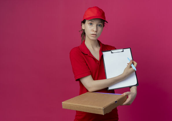 印象令人印象深刻的年轻漂亮的送货女孩穿着红色制服 戴着帽子拿着比萨饼包和剪贴板 用钢笔指着剪贴板 在深红色的背景上与复制空间隔离帽子持有钢笔
