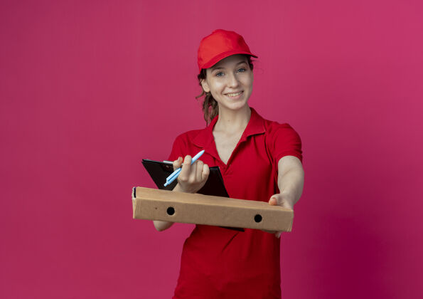 披萨微笑着的年轻漂亮的送货女孩 穿着红色制服 戴着帽子 对着镜头伸出披萨包 手里拿着一支笔 剪贴板被隔离在深红色的背景上 还有复印空间拿着微笑剪贴板