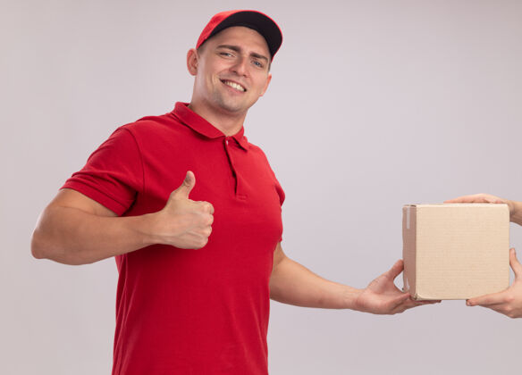 表演面带微笑的年轻送货员穿着制服 拿着送帽盒 站在白墙上向客户竖起大拇指人表情递送