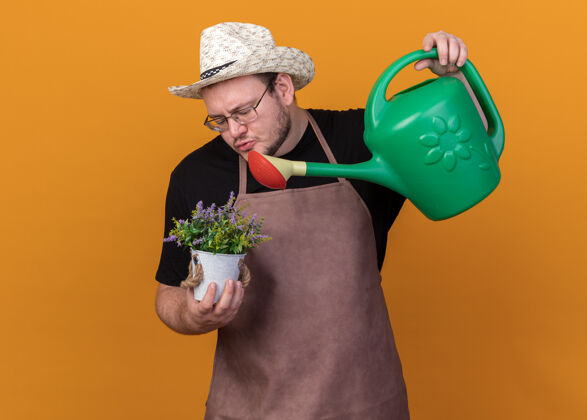 感觉高兴的年轻男园丁戴着园艺帽和手套在花盆里浇花用浇水罐隔离在橙色的墙上站男脸