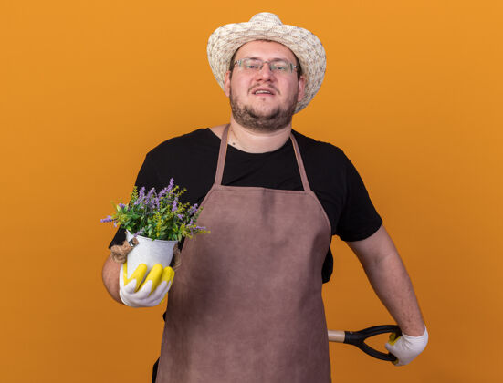 脸自信的年轻男园丁戴着园艺帽和手套 腰间拿着铲子 花盆里的花被隔离在橙色的墙上人穿年轻