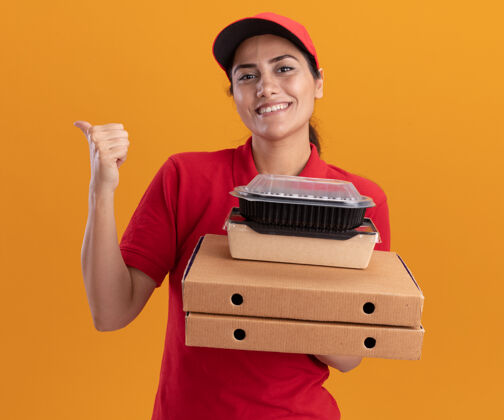 表情微笑着的年轻送货女孩穿着制服 戴着帽子 手里拿着披萨盒和食品容器 后面是橙色的隔离墙 有复印空间拿着披萨帽子