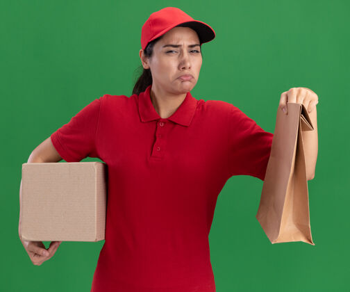 盒子悲伤的年轻送货女孩穿着制服和帽子与纸食品包装盒绿色隔离墙举行表情人制服