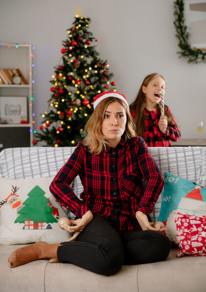 享受快乐的女儿吃着糖果棒 愤怒的母亲拿着玻璃球饰品坐在沙发上 在家里享受圣诞节时光女儿坐着球