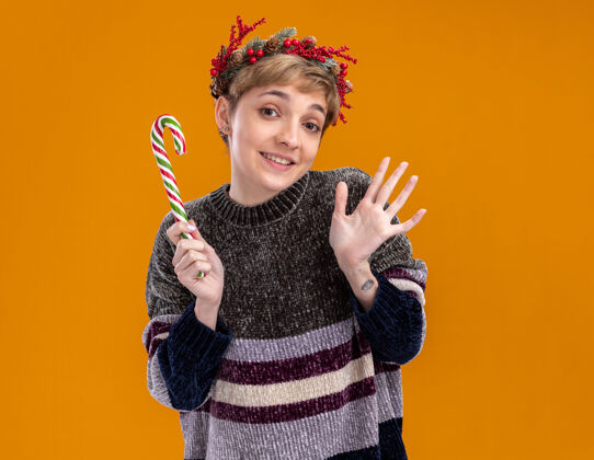 表演快乐的年轻漂亮女孩戴着圣诞花环手持圣诞甜手杖看着相机显示空手孤立在橙色的背景手杖头手