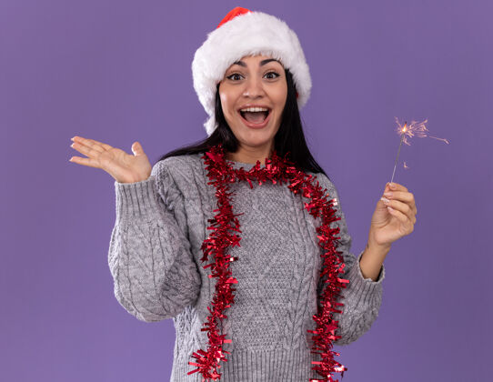 年轻令人印象深刻的年轻白人女孩戴着圣诞帽 脖子上戴着金箔花环 手里拿着节日火花灯 看着镜头 在紫色背景上孤立着一只空手花环印象圣诞节