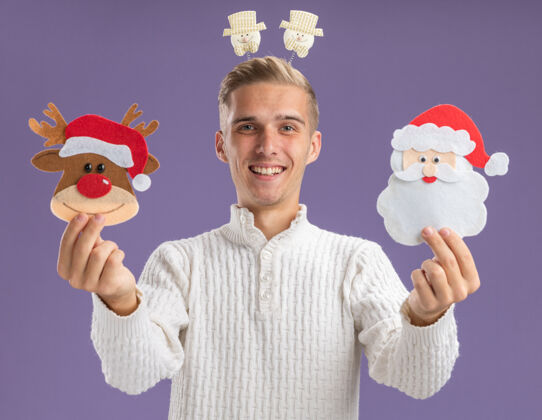 圣诞快乐的年轻帅哥戴着雪人头带 向镜头伸出圣诞纸饰品 看着隔离在紫色背景上的镜头头带伸展年轻