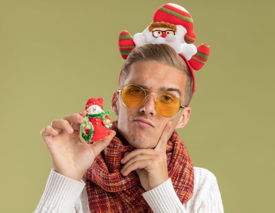 围巾可疑的年轻帅哥戴着圣诞老人的头带和围巾看着相机拿着雪人圣诞饰品手指贴在脸上橄榄绿的背景隔离圣诞雪人年轻