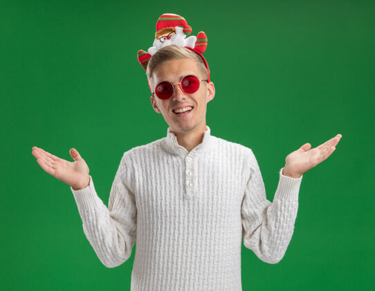 圣诞老人快乐的年轻帅哥戴着圣诞老人的头带 戴着眼镜 看着镜头 两手空空的 孤立在绿色的背景上格林圣诞老人头带