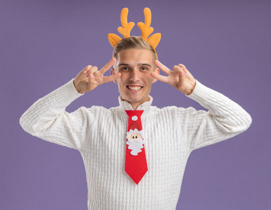 家伙快乐的年轻帅哥戴着驯鹿的鹿角头带和圣诞老人的领带 看着摄像机 在紫色背景上孤立的眼睛附近显示v符号圣诞老人象征Vsign