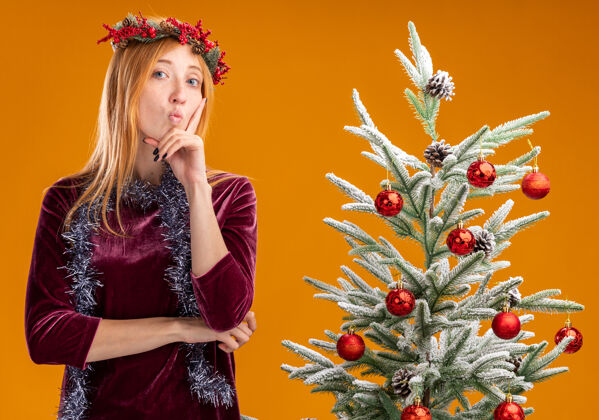 手指令人印象深刻的年轻美丽的女孩站在圣诞树附近穿着红色的裙子和花环与花环的脖子上把手指放在脸颊上孤立的橙色背景印象年轻圣诞树