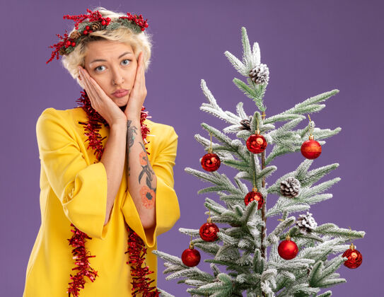 年轻无聊的金发女郎头戴圣诞花环 脖子上戴着金属丝花环 站在装饰好的圣诞树旁 看着摄像机 手放在脸上 在紫色背景下与世隔绝圣诞树花环花圈