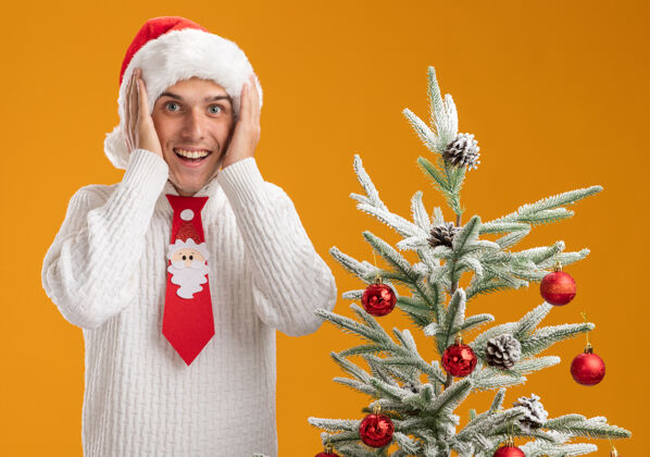 圣诞树令人印象深刻的年轻帅哥戴着圣诞帽 打着圣诞老人的领带 站在装饰好的圣诞树旁 手放在头上 看着橙色背景上的相机帽子站手