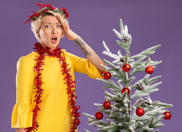 紫色戴着圣诞花环 脖子上戴着金属丝花环的年轻金发女人站在装饰好的圣诞树旁 看着相机 手放在头上 在紫色的背景下与世隔绝圣诞快乐关心年轻