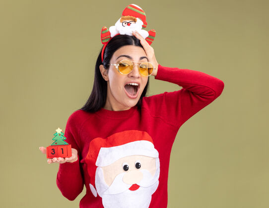 圣诞老人令人印象深刻的年轻白人女孩戴着圣诞老人的头带和毛衣戴着眼镜拿着圣诞树玩具和日期看着相机保持手在头上橄榄绿的背景隔离头带举行手