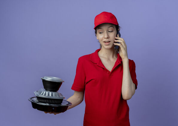 制服令人印象深刻的年轻漂亮的送货女孩穿着红色制服 戴着帽子 看着食品容器 在紫色背景上与复印空间隔离的电话交谈漂亮食物紫色