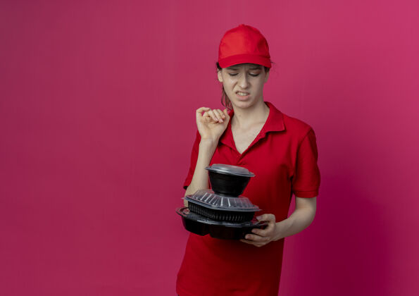 制服恼怒的年轻漂亮的送货女孩穿着红色制服 戴着帽子 看着被隔离在深红色背景下的食品容器 还有复印空间交货容器食物