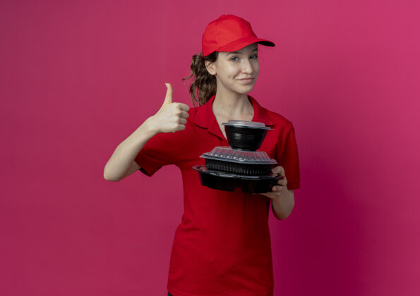 向上令人高兴的年轻漂亮的送货女孩穿着红色制服 戴着帽子拿着食物容器 在深红色背景上孤立地竖起大拇指 留着复印空间拇指帽子女孩