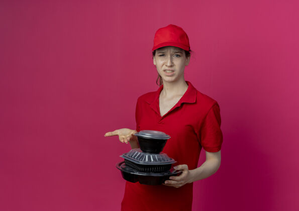 红色年轻漂亮的送货女孩穿着红色制服 戴着帽子 手持并用手指着隔离在深红色背景上的食品容器 还有复印空间手持有年轻