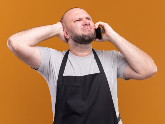 手一个穿着制服的中年男性理发师正在打电话 把手放在隔离在橙色墙上的头后面理发师站电话