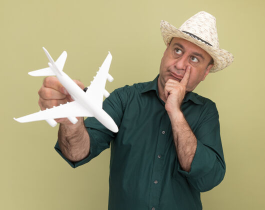 思想想着看着身边的中年男子穿着绿色t恤 戴着帽子拿着玩具飞机把手放在脸颊上隔离在橄榄绿的墙上飞机穿着玩具