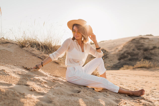 旅行在沙漠沙滩上穿着白色衣服戴着草帽的时髦美女日落太阳镜女士女性