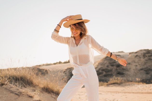 狩猎时尚快乐的美丽微笑的女人在沙漠里摆姿势 穿着白色的衣服 戴着草帽和太阳镜在夕阳下人非洲休闲