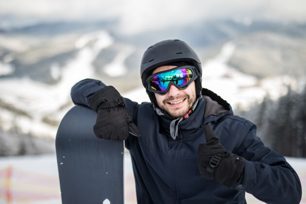 人在冬季滑雪场 站在雪坡顶上的男子滑雪板手拿着滑雪板 对着镜头微笑着竖起大拇指肖像雪头