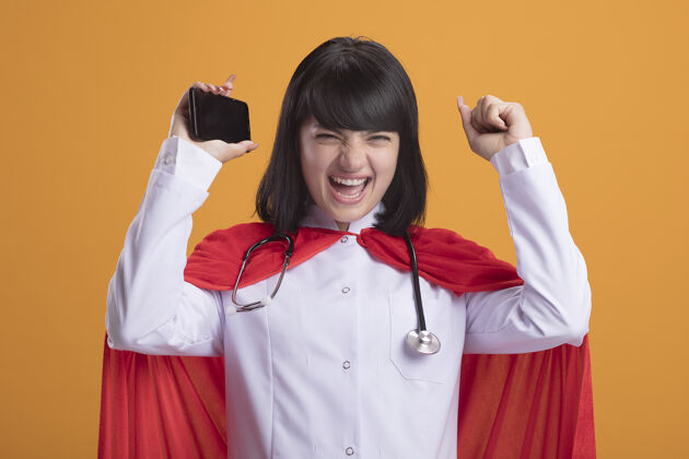 医疗兴奋的年轻超级英雄女孩戴着听诊器 穿着医用长袍和斗篷 手里拿着电话 举手兴奋人抱着