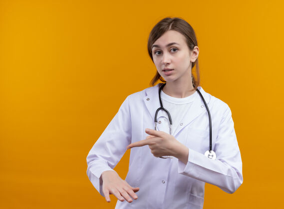 复制焦急的年轻女医生穿着医用长袍 用听诊器指着她手上孤立的橙色空间和复印空间年轻医生指向
