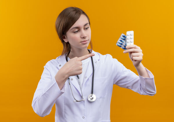 医生自信的年轻女医生 穿着医用长袍 手持听诊器 拿着医用药品 指着孤立的橙色空间持有信心医疗