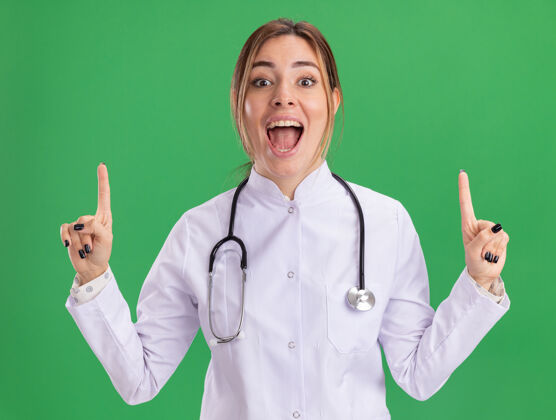 医学惊讶的年轻女医生穿着医用长袍 用听诊器指着隔离在绿色墙上的那堵墙女性表情站