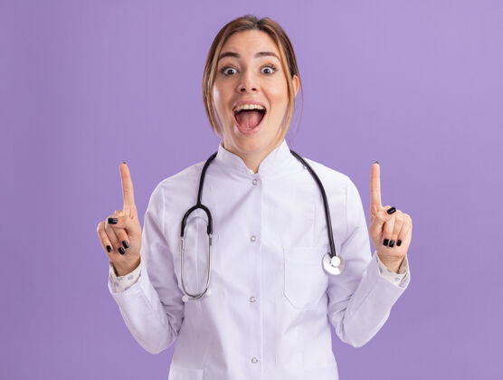 感情印象深刻的年轻女医生穿着医用长袍 用听诊器指着隔离在紫色墙上的复印空间人衣服立场