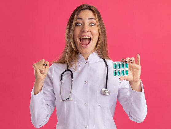 兴奋兴奋的年轻女医生穿着医用长袍 手持听诊器的药片 在粉红色的墙上显示“是”的手势表演药丸抱着