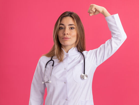 人自信的年轻女医生穿着医用长袍 听诊器显示出强烈的手势隔离在粉红色的墙上表演自信表情