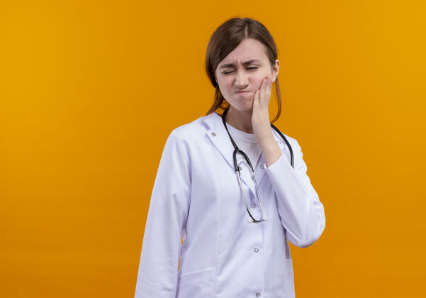 磨损疼痛的年轻女医生穿着医用长袍和听诊器 手放在脸颊上 忍受着牙痛 在隔离的橙色空间和复印空间太空脸颊医生