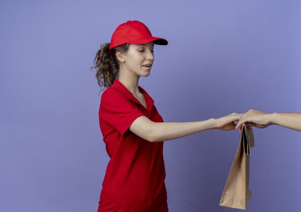 漂亮令人印象深刻的年轻漂亮的送货女孩穿着红色制服 戴着帽子站在侧视图给纸包给客户隔离在紫色背景与复印空间背景给予印象