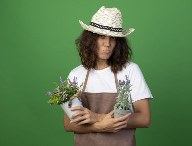 人穿着制服的年轻女园丁戴着园艺帽 在花盆里拿着花和交叉女人成人穿