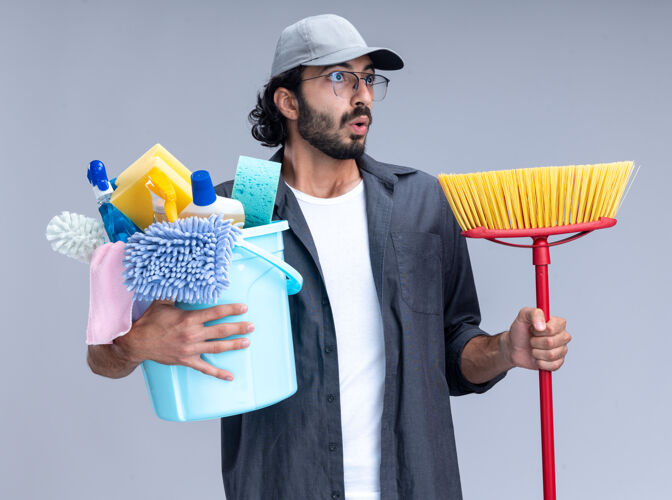 帅哥惊讶的看着身边年轻帅气的清洁工穿着t恤 戴着帽子 拿着一桶清洁工具 用拖把隔离在白色的墙上穿举行清洁