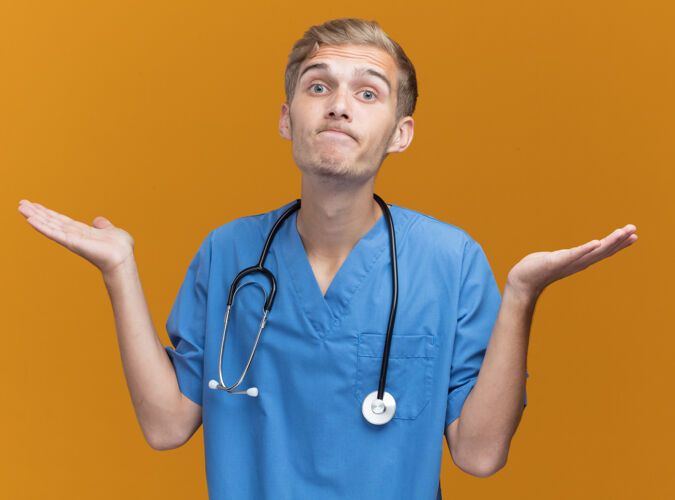 男性困惑的年轻男医生穿着医生制服听诊器摊开双手隔离在橙色的墙上年轻手制服