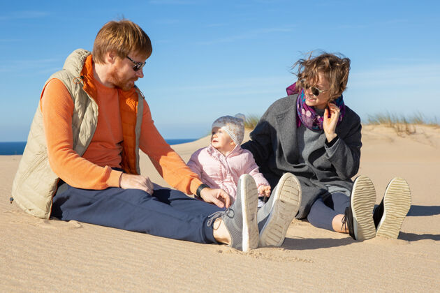 海边快乐的父母和可爱的女婴穿着暖和的衣服 享受海上的休闲时光 一起坐在沙滩上关怀夫妻家庭