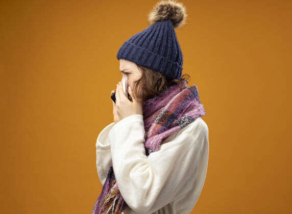 长袍站在侧视图年轻的生病的女孩穿着白色长袍 戴着冬天的帽子和围巾讲电话孤立在橙色说话穿年轻