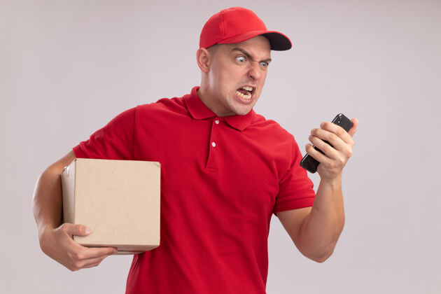 年轻愤怒的年轻送货员穿着制服 戴着帽子 手里拿着盒子 看着手机孤立在白墙上送货帽子手