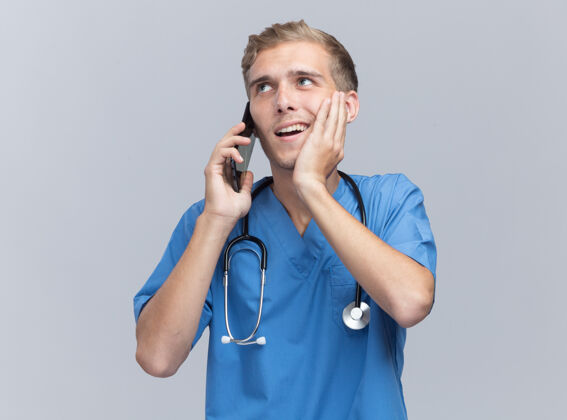 面颊面带微笑的年轻男医生穿着医生制服 手持听诊器讲电话 手放在隔离在白墙上的脸颊上微笑年轻医生