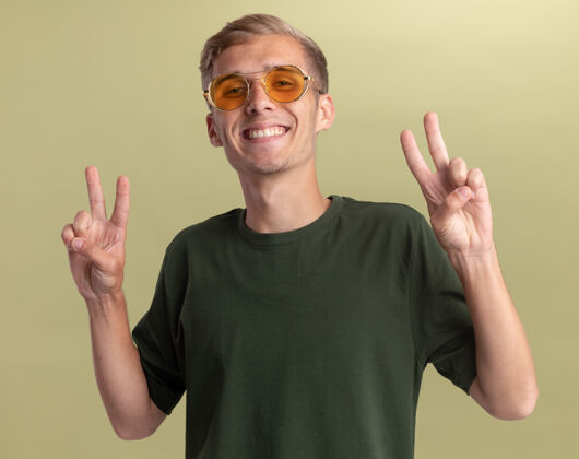 眼镜微笑着的年轻帅哥穿着绿色衬衫 戴着眼镜 在橄榄绿的墙上显示出与世隔绝的和平姿态秀男人橄榄