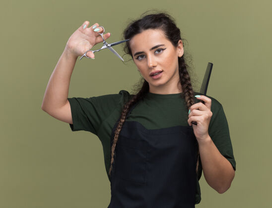 女高兴的年轻女理发师在统一举行梳子和提高剪子隔离在橄榄绿的墙上橄榄色梳子年轻